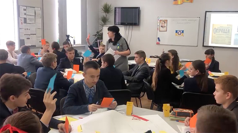 A teacher and students in a classroom in Ukraine. The students are doing a media literacy exercise with orange and blue pieces of paper.