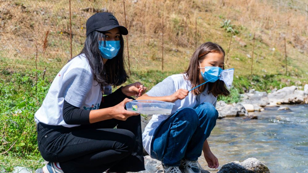 Photo of two girls squatting by the river side collecting samples from the water. The girls are wearing masks.