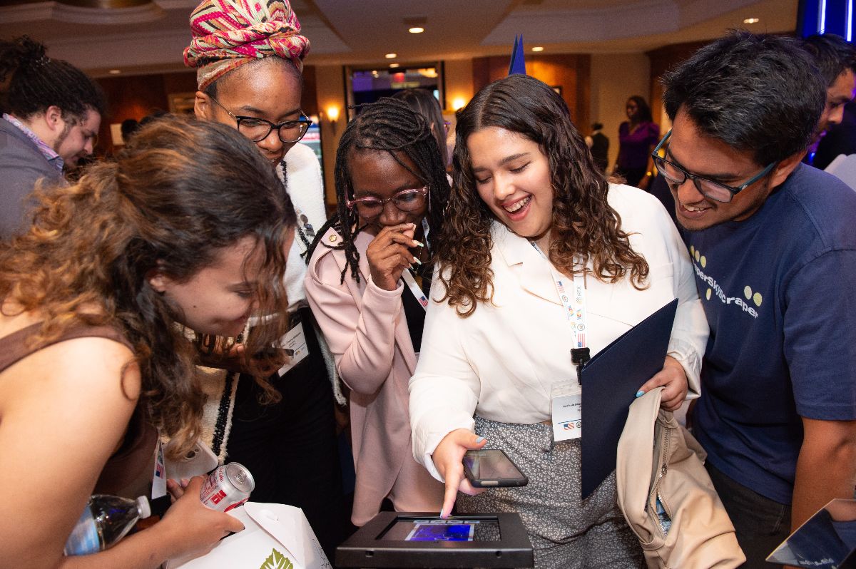 A group of youth are standing in a group smiling at something they are viewing on a device 