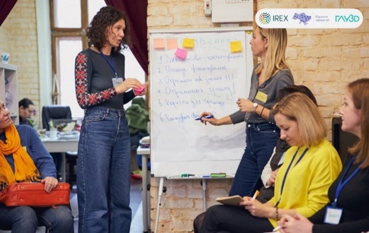 Image of two women stading next to flip chart and taking notes, they are surrounded by other women who appear to be in a training 